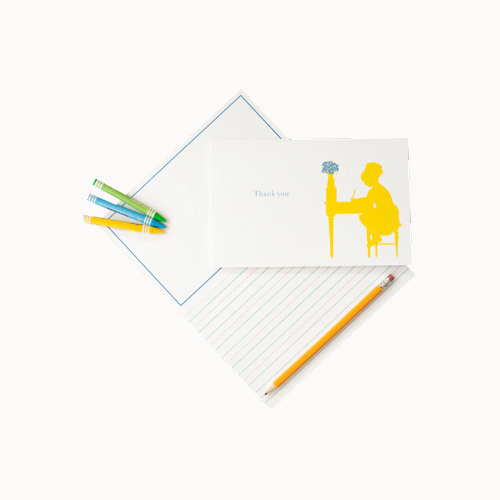 Thank you card with yellow illustration of a curly-haired girl writing at her desk, surrounded by another open gift card and three crayons