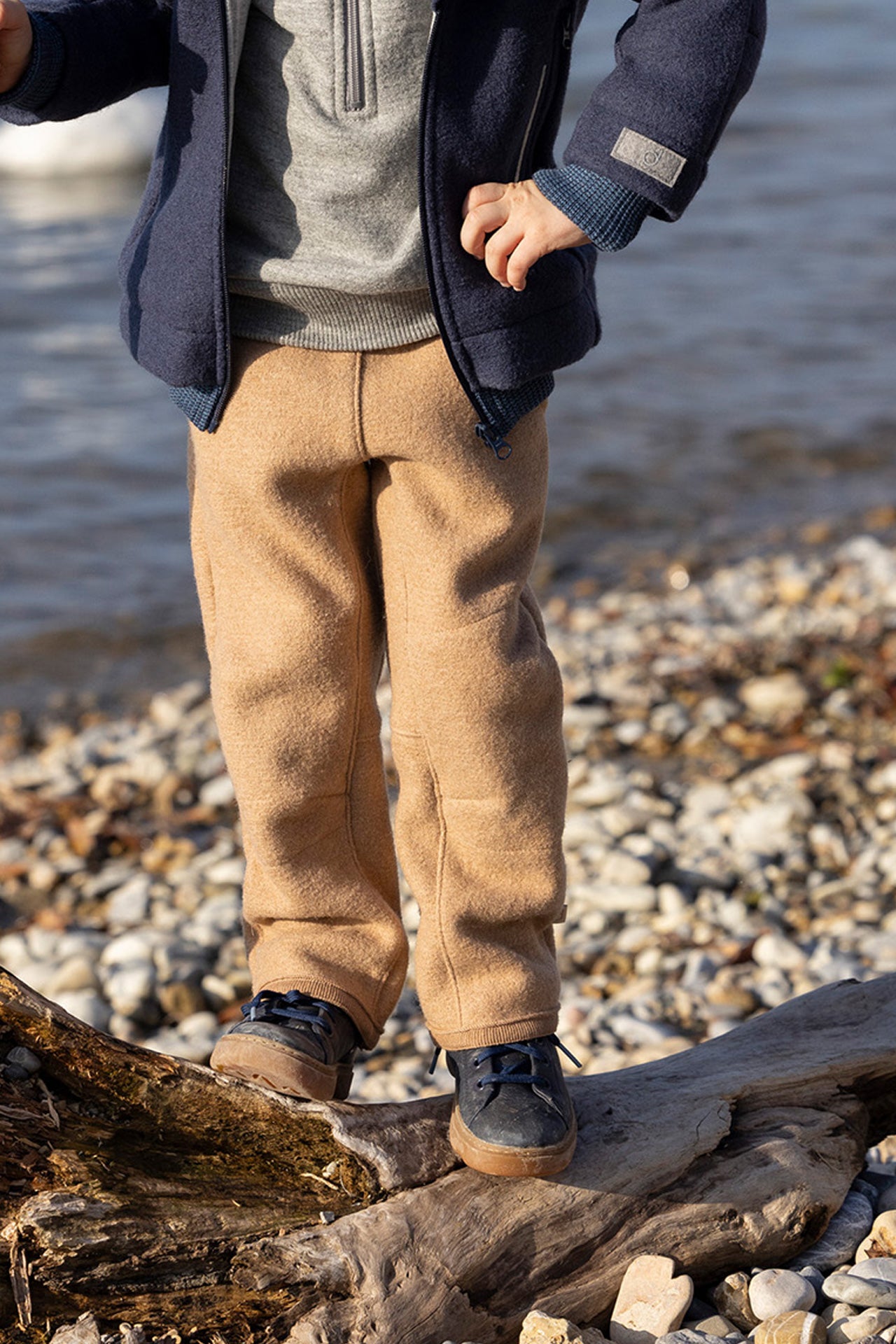 Person wearing beige pants and navy coat standing on a pebbly beach.