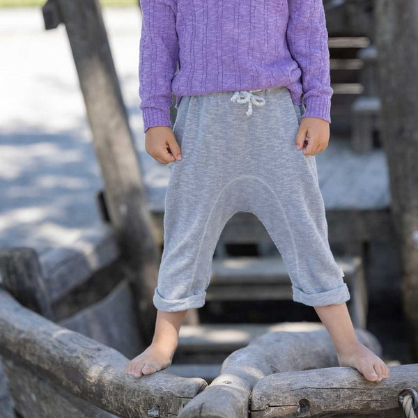 A child standing on a log by the water wearing grey knitted linen trousers with a soft elastic waistband and rolled-up legs, with knitted back pockets.