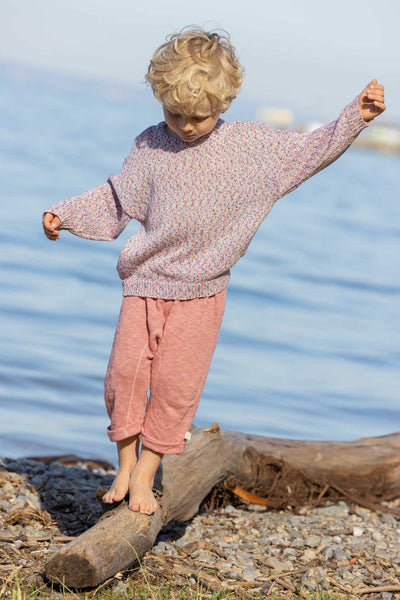 A child standing on a log by the water wearing pink knitted linen trousers with a soft elastic waistband and rolled-up legs, with knitted back pockets.