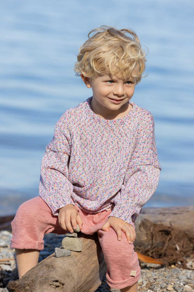 A child sitting on a log by the water wearing pink knitted linen trousers with a soft elastic waistband and rolled-up legs, with knitted back pockets.