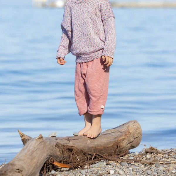 A child standing on a log by the water wearing pink knitted linen trousers with a soft elastic waistband and rolled-up legs, with knitted back pockets.