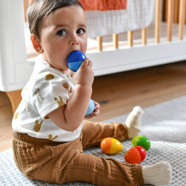 Baby sitting on a rug holding a blue teething toy with colorful toys around