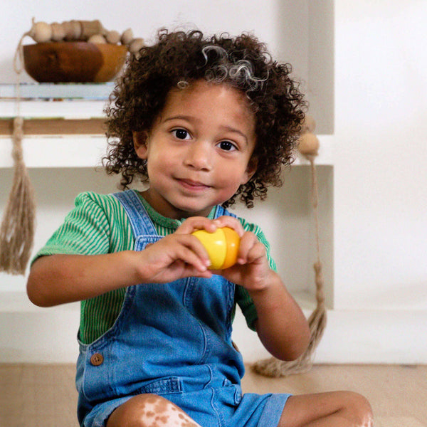 Child holding a yellow ball in a room with shelves and toys.
