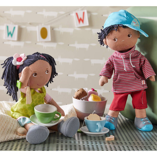 Two dolls sitting at a tea party with cups and saucers on a table.