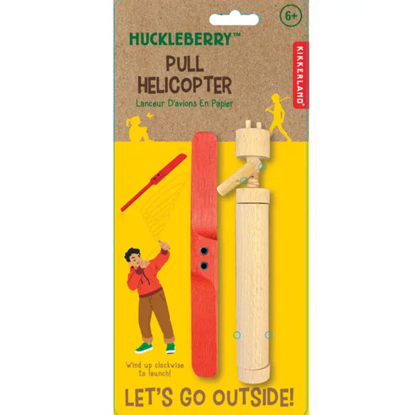 Packaged Huckleberry Pull Helicopter toy designed by Koens & Middlekoop, made from wood and polyester string, with a red launcher, viewed through a yellow die-cut window on the packaging, with 'LET'S GO OUTSIDE!' text.
