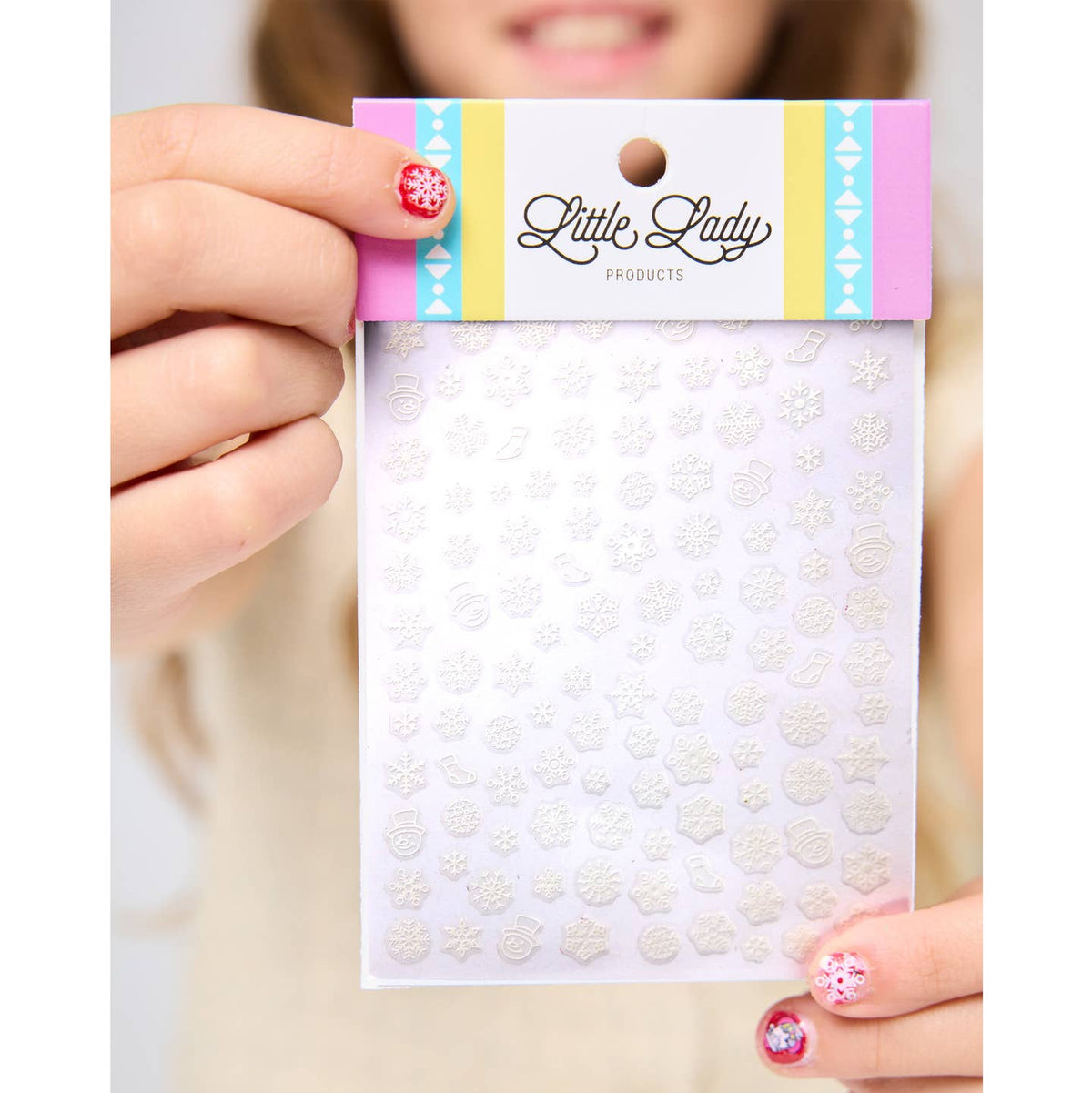 A person holds a package of Little Lady Products white snowflake nail stickers. The packaging features a pastel pink, yellow, and blue header card with the brand name 'Little Lady Products' printed in black script.