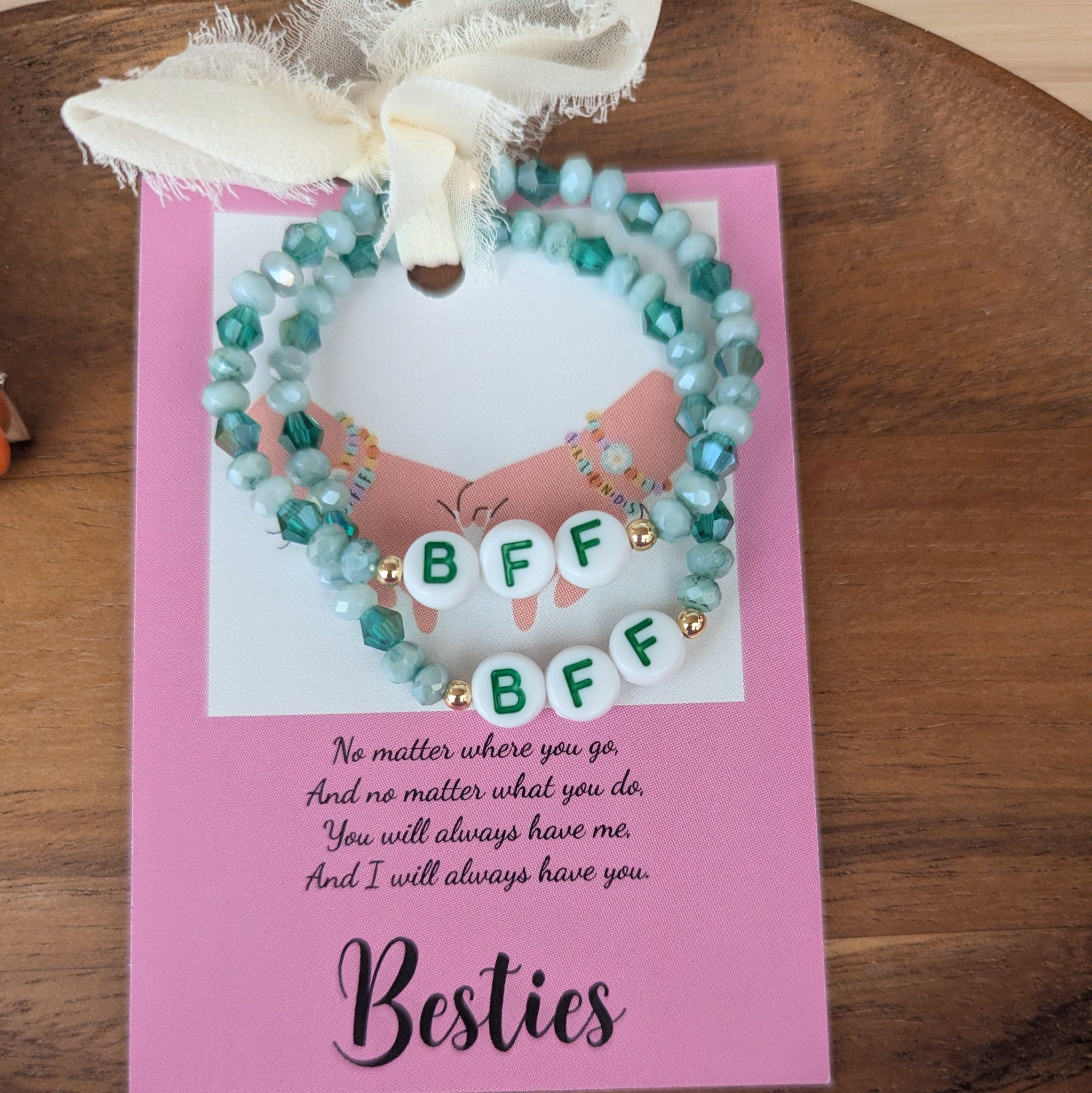 Two beaded bracelets on a pink card with 'BFF' text, tied with a white ribbon on a wooden surface.