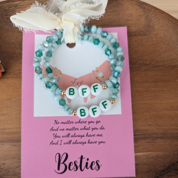 Two beaded bracelets on a pink card with 'BFF' text, tied with a white ribbon on a wooden surface.