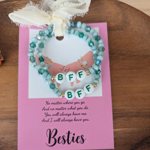 Two beaded bracelets on a pink card with 'BFF' text, tied with a white ribbon on a wooden surface.