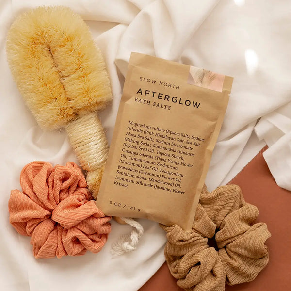 Bath salts package with a loofah, scrubber, and scrunchie on a soft fabric background