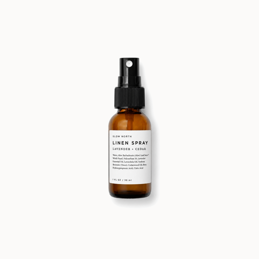 A small amber glass spray bottle with a black pump and a white label reading 'LINEN SPRAY' and 'LAVENDER + CEDAR', centered on a white background.