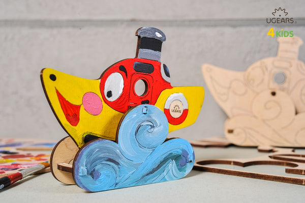Colorful toy steamboat with a red mouth and blue waves on a gray background, featuring UGears Kids branding.