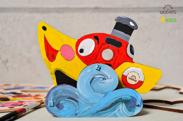 Colorful toy steamboat with a red mouth and blue waves on a gray background, featuring UGears Kids branding.