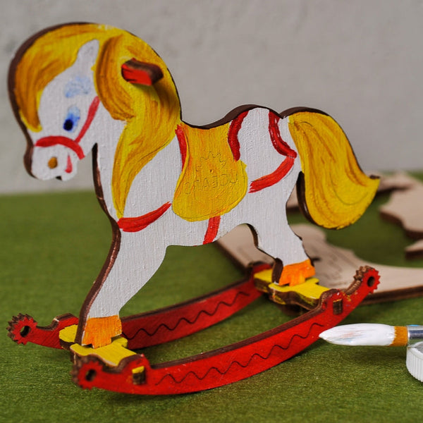 Wooden horse model with paintbrush and paint on a green surface, UGEARS brand visible