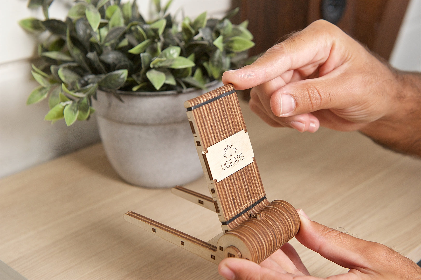 Wooden model of a phone holder with a brand label, held by a person on a wooden surface with a plant in the background.