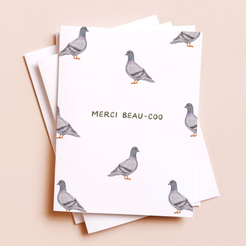 A stack of white greeting cards printed with watercolor pigeon illustrations and the French text 'MERCI BEAU-COO' in the center, photographed on a soft peach background.
