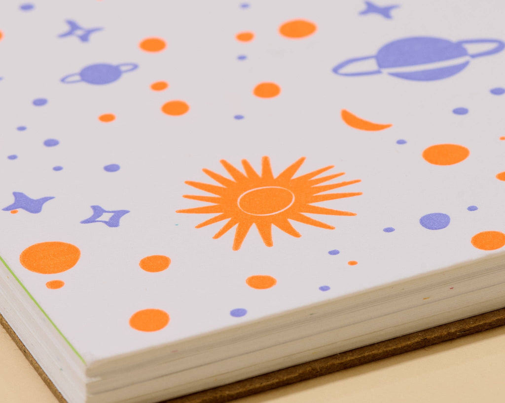 A close-up of a notebook or journal with a white cover decorated with colorful space-themed illustrations, including an orange sun, blue planets, stars, and dots.