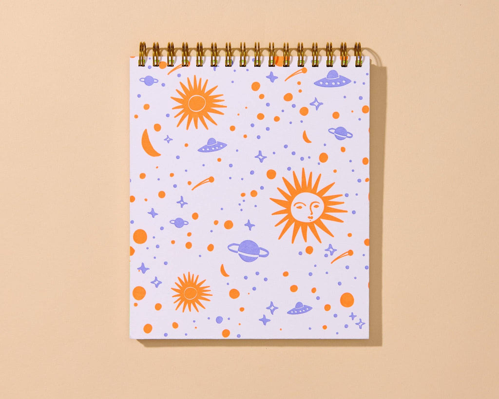 A spiral-bound notebook with a white cover decorated with a colorful space-themed pattern of orange suns, purple planets and moons, and small stars, shown on a warm beige background.