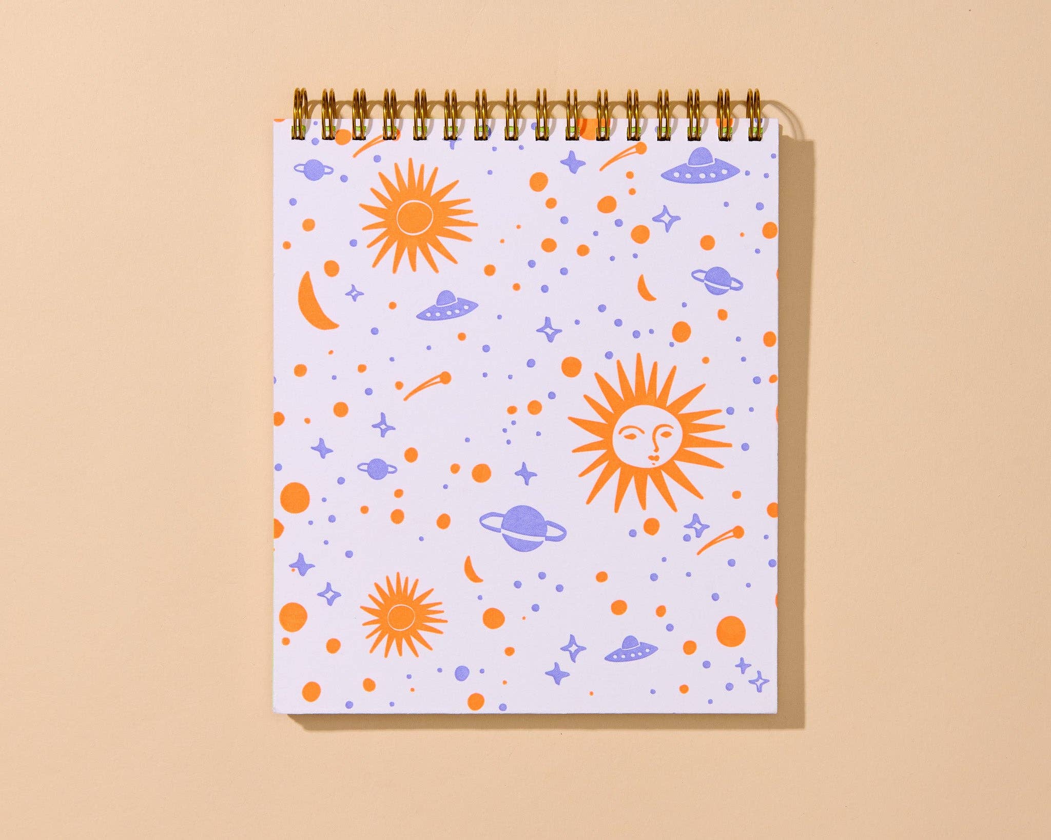 A spiral-bound notebook with a white cover decorated with a colorful space-themed pattern of orange suns, purple planets and moons, and small stars, shown on a warm beige background.