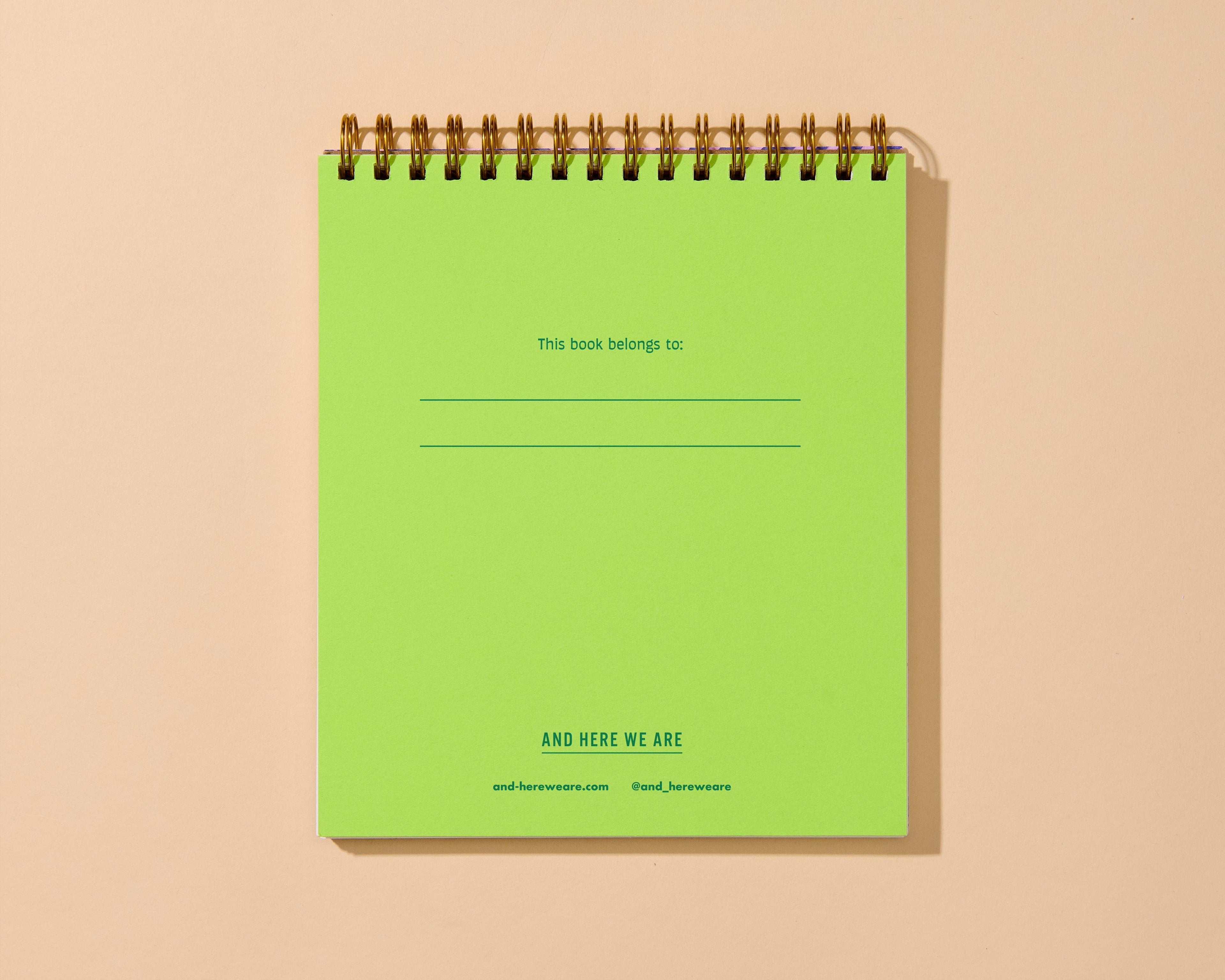 A lime green spiral-bound notebook with gold-toned metal wire binding, shown standing upright against a warm beige background. The cover has printed text including 'This book belongs to:' with blank lines and 'AND HERE WE ARE' near the bottom.
