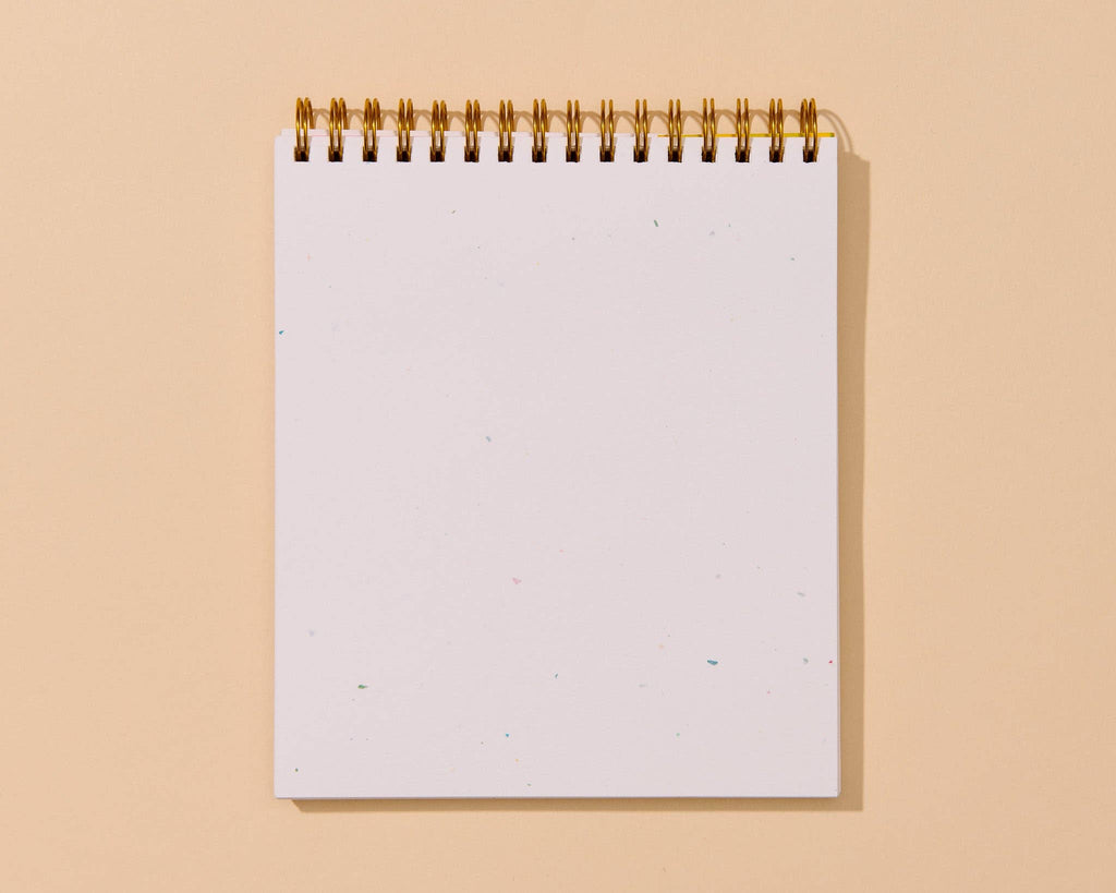 A blank white spiral-bound notepad with a gold wire binding, standing upright against a solid warm beige background.