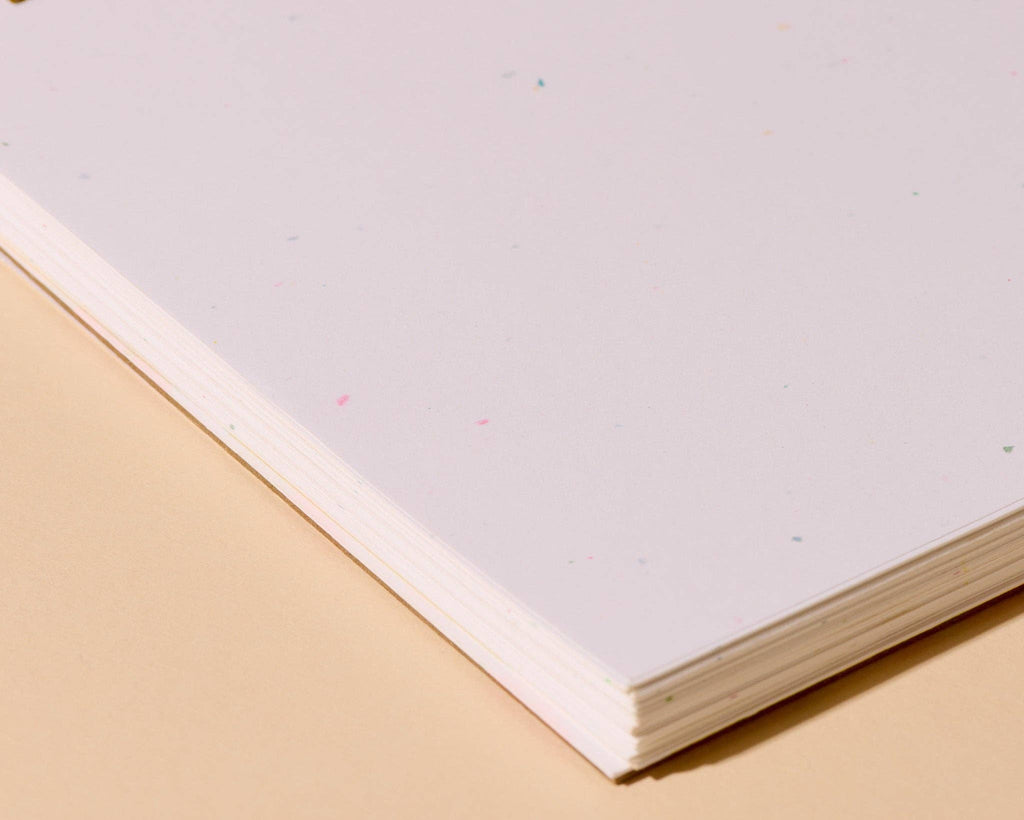 A close-up of a stack of white paper sheets with tiny multicolored flecks, resting on a warm beige surface.