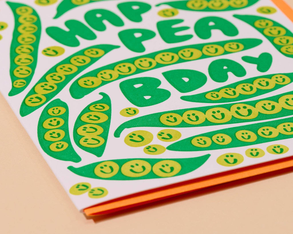 A close-up of a printed card or paper design with the words “HAPPENING PEANUT DAY” in bold green letters, decorated with green pea pods and yellow smiley faces on a white background, with orange and peach layers visible along the edge.
