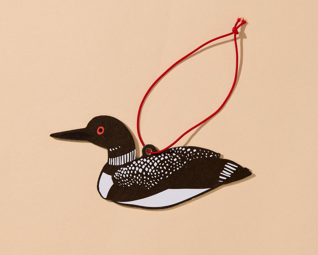 A paper cutout loon ornament with a red hanging string, shown on a solid beige background.