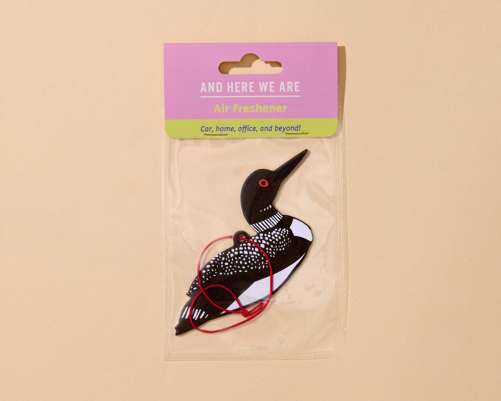 A loon-shaped air freshener in a clear plastic bag with a pink and green header card that reads 'AND HERE WE ARE' and 'Air freshener'. The loon is black and white speckled with a red eye and red string nozzle.