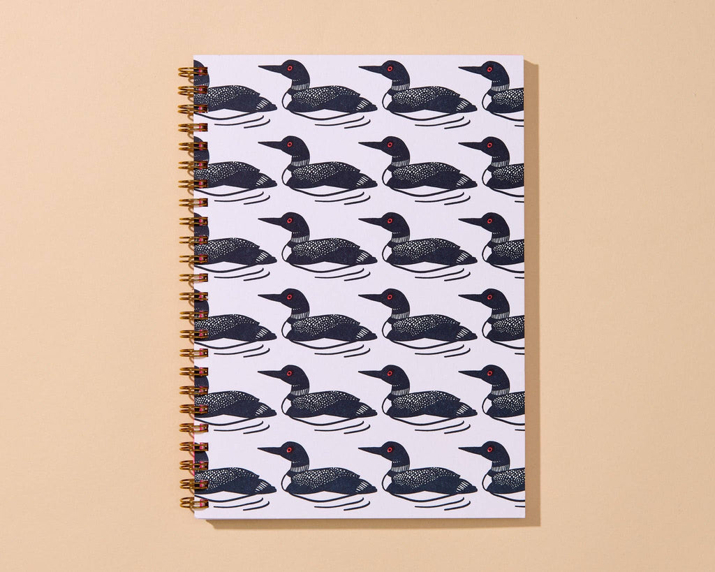A spiral-bound notebook with a repeating pattern of black and white loons on a white cover, set against a solid beige background.