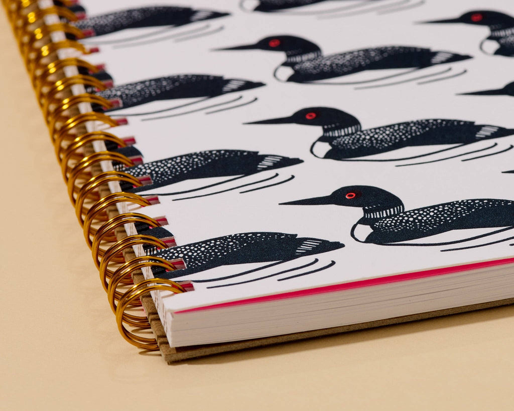 A close-up of a spiral-bound notebook with a white cover printed with black loon illustrations and small red eye accents, bound with a gold spiral on a beige surface.