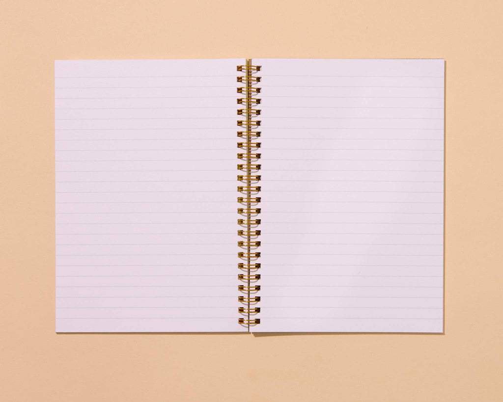An open lined spiral notebook with gold-toned wire binding, centered on a warm beige background.