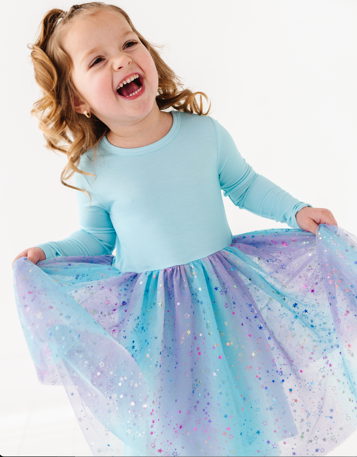 A young girl with blonde hair in pigtails is smiling and laughing while twirling, wearing a light blue long-sleeve top and a sparkly blue, purple, and pink tulle tutu skirt against a plain white background.
