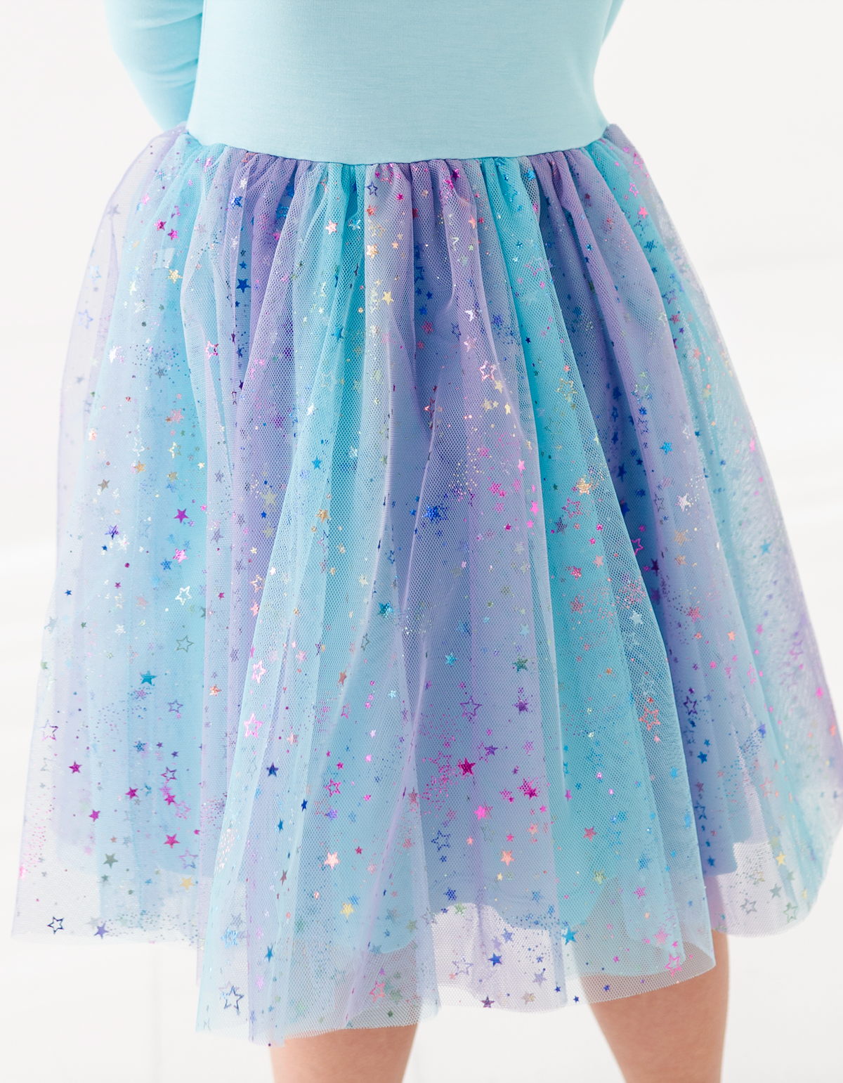 A close-up of a child wearing a voluminous tulle tutu skirt in pastel blue, lavender, and purple with scattered multicolored star glitter, paired with a light blue top against a white background.