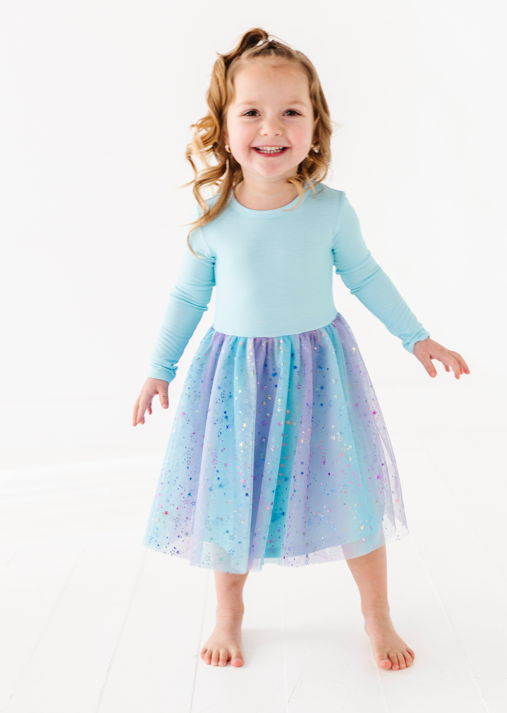A young girl with blonde hair in a ponytail stands barefoot on a white studio floor, wearing a light blue long-sleeve top and a sparkly tulle skirt that fades from blue to lavender.