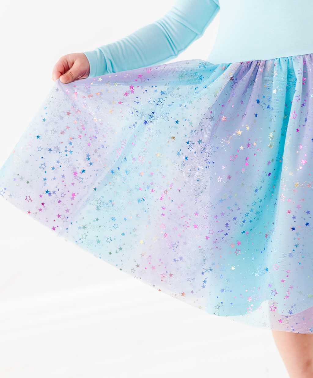A close-up of a child wearing a light blue long-sleeve top and a flared tulle skirt with a pastel gradient of blue, purple, and pink, decorated with scattered iridescent star glitter.