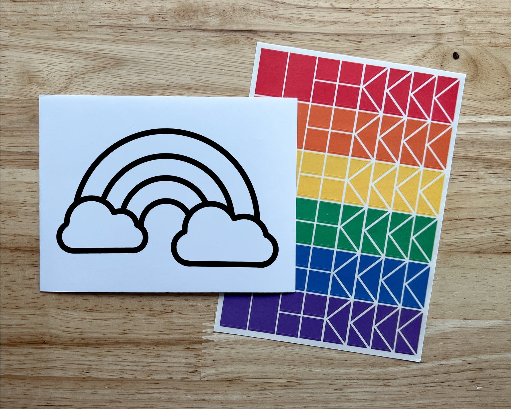 Paper with rainbow and cloud design and a sheet of geometric patterned stickers on a wooden surface.