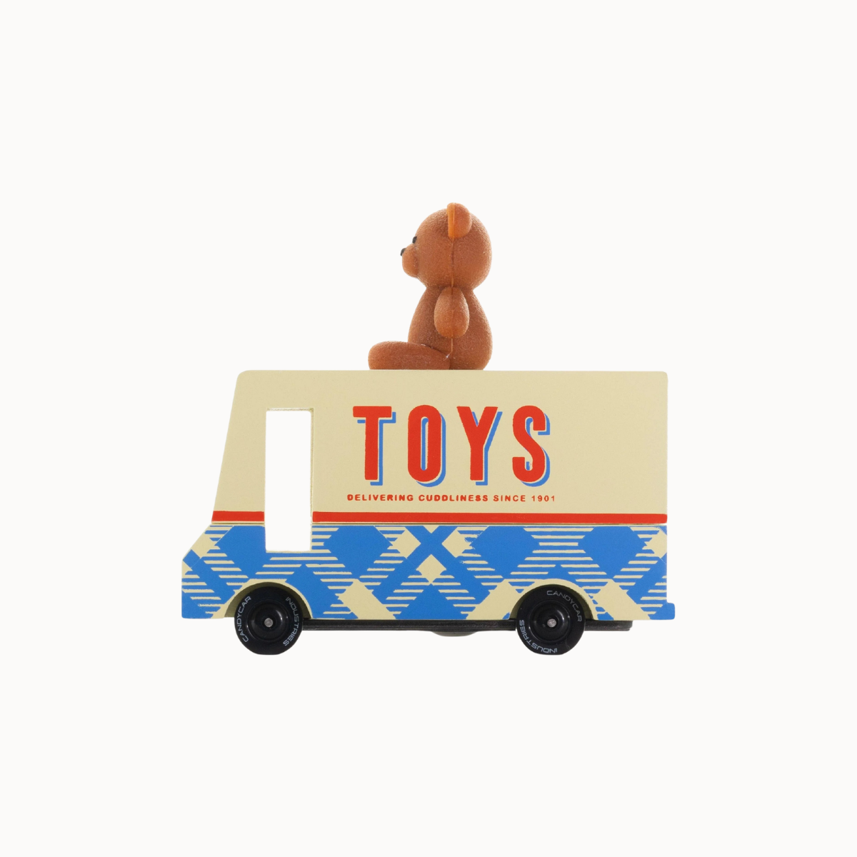 A wooden toy delivery truck with a small brown teddy bear sitting on top. The truck has a cream-colored upper body with 'TOYS' written in red and blue letters, and a blue and cream geometric pattern on the lower half.