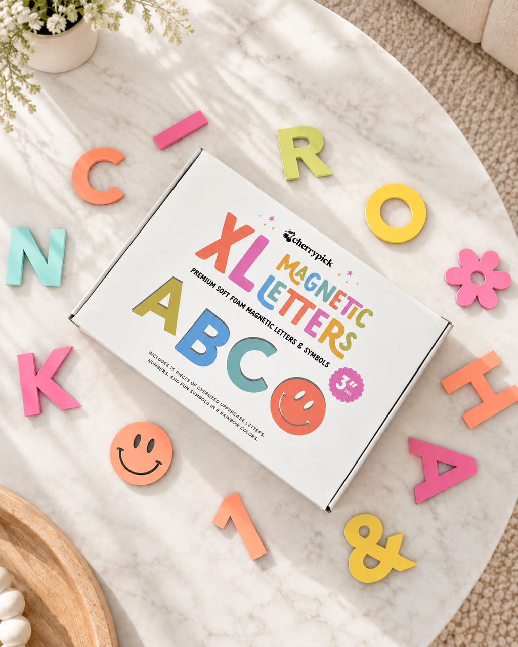 A white box labeled 'XL Magnetic Letters' is placed on a light marble surface, surrounded by colorful magnetic letters and smiley faces. The scene is styled as a flat lay with soft natural shadows.