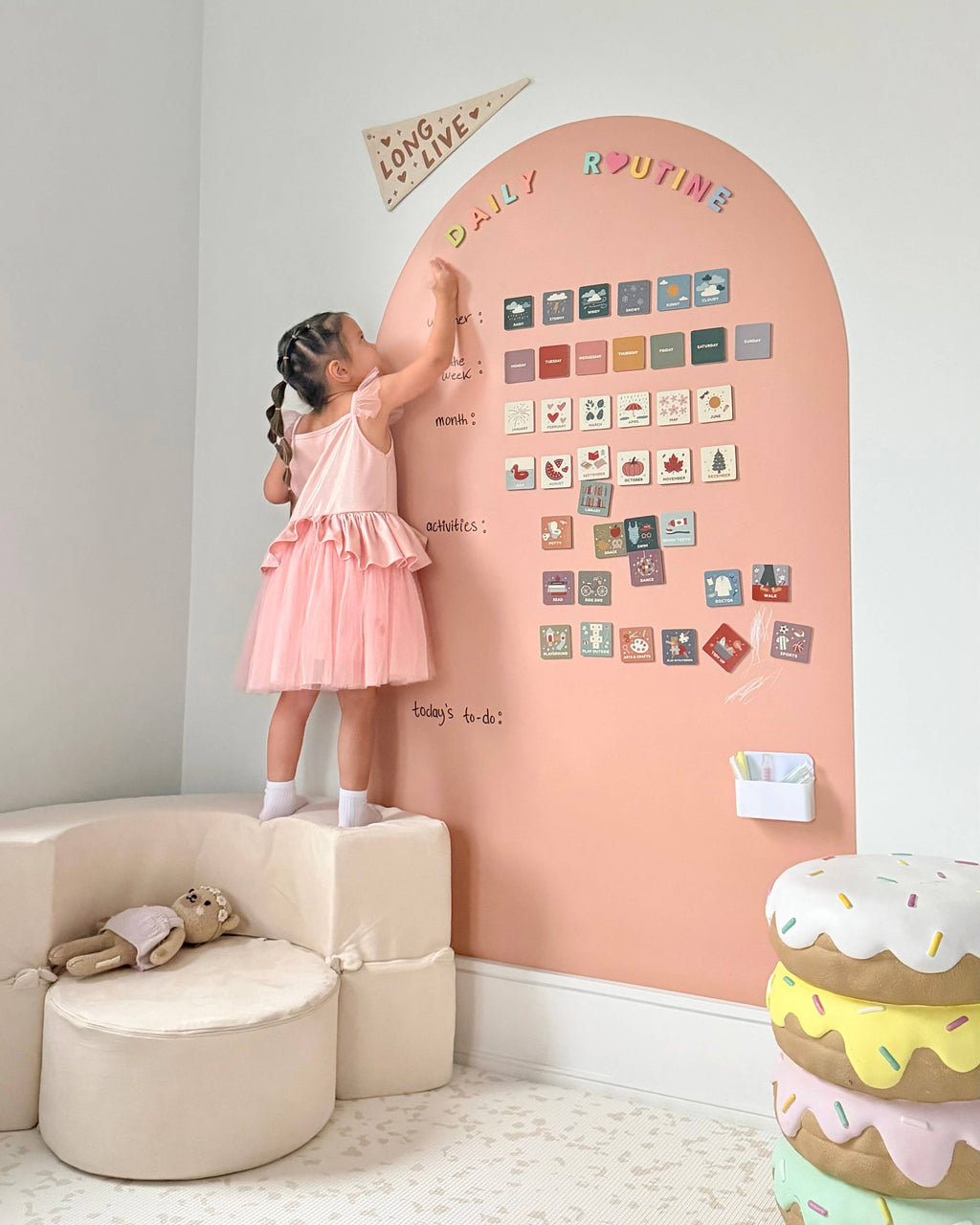 Child in a pink dress standing on a small stool, reaching to place a magnet on a pink board with daily routine chart.
