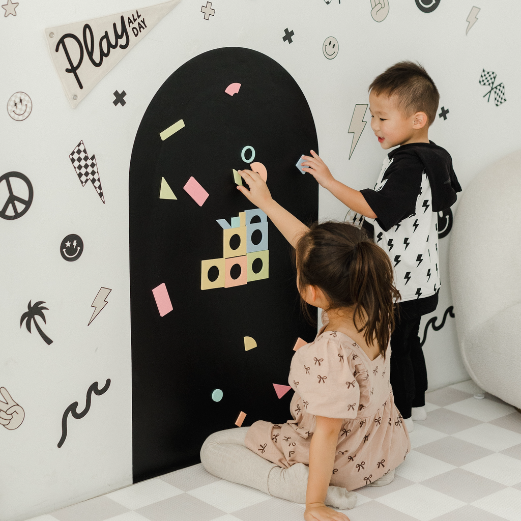 Two children playing with colorful shapes on a blackboard wall in a room with decorative wall stickers.