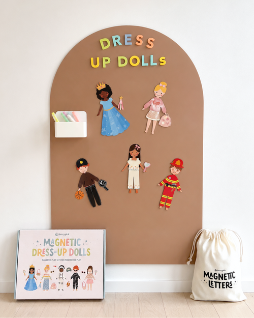 Dress-up dolls set with magnetic letters on a brown board, next to a box and drawstring bag.