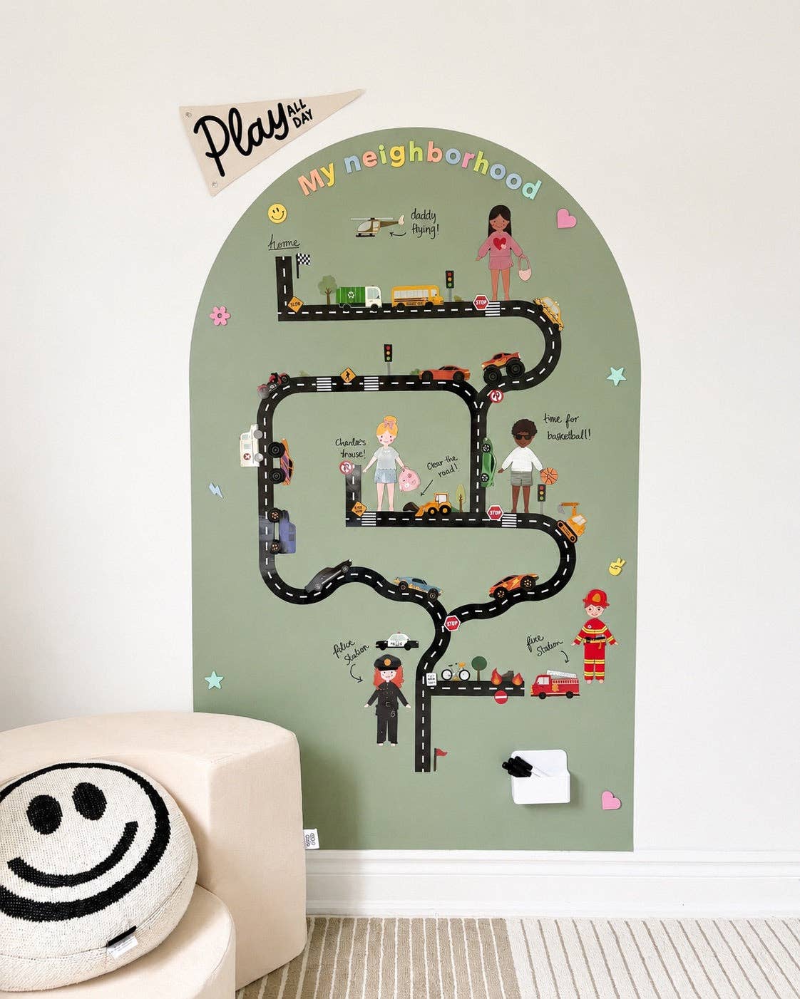 A children's play neighborhood wall mural featuring a winding black road with toy cars, illustrated characters, and colorful text including 'My neighborhood' and 'Play with me,' shown mounted on a white wall above a cream chair.