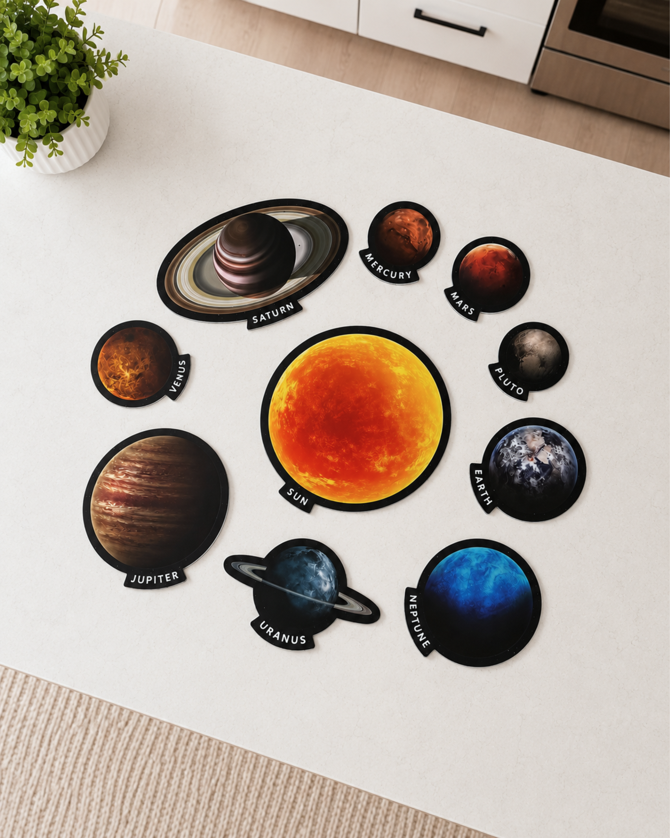 A circular solar system wall decal set displayed on a white wall, featuring a large yellow sun in the center with eight planets orbiting it, each labeled with its name. The scene is photographed from above in a bright room with a small potted plant in the top-left corner.