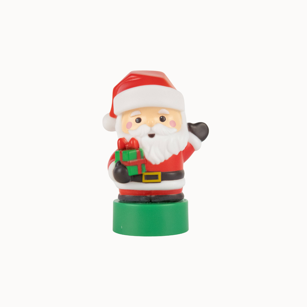 A small Santa Claus figurine standing on a round green base, wearing a red hat and coat with a white beard, and holding a green gift box with a red bow. The figurine is photographed against a plain light gray background.