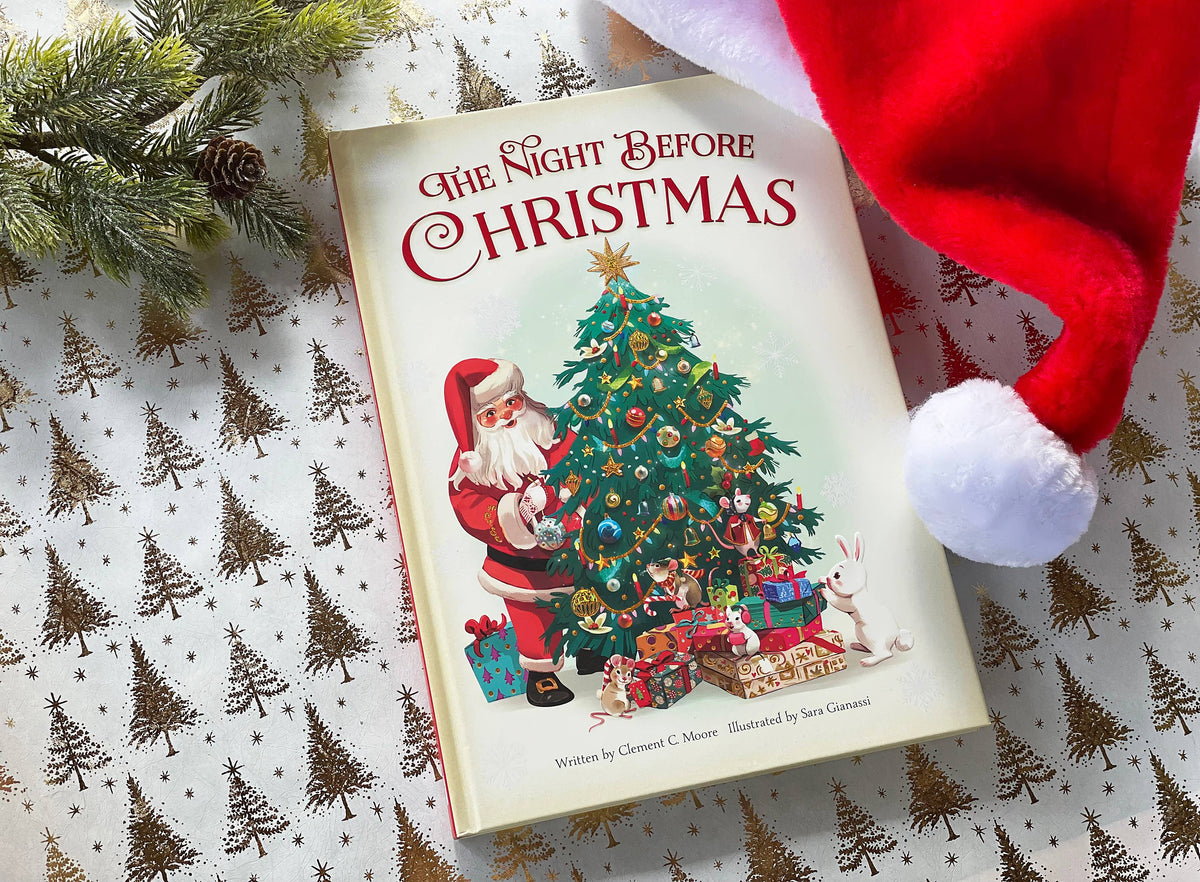A children's book titled "The Night Before Christmas" lies on a patterned surface, surrounded by a red Santa hat and evergreen branches. The cover shows Santa Claus beside a decorated Christmas tree with wrapped gifts.
