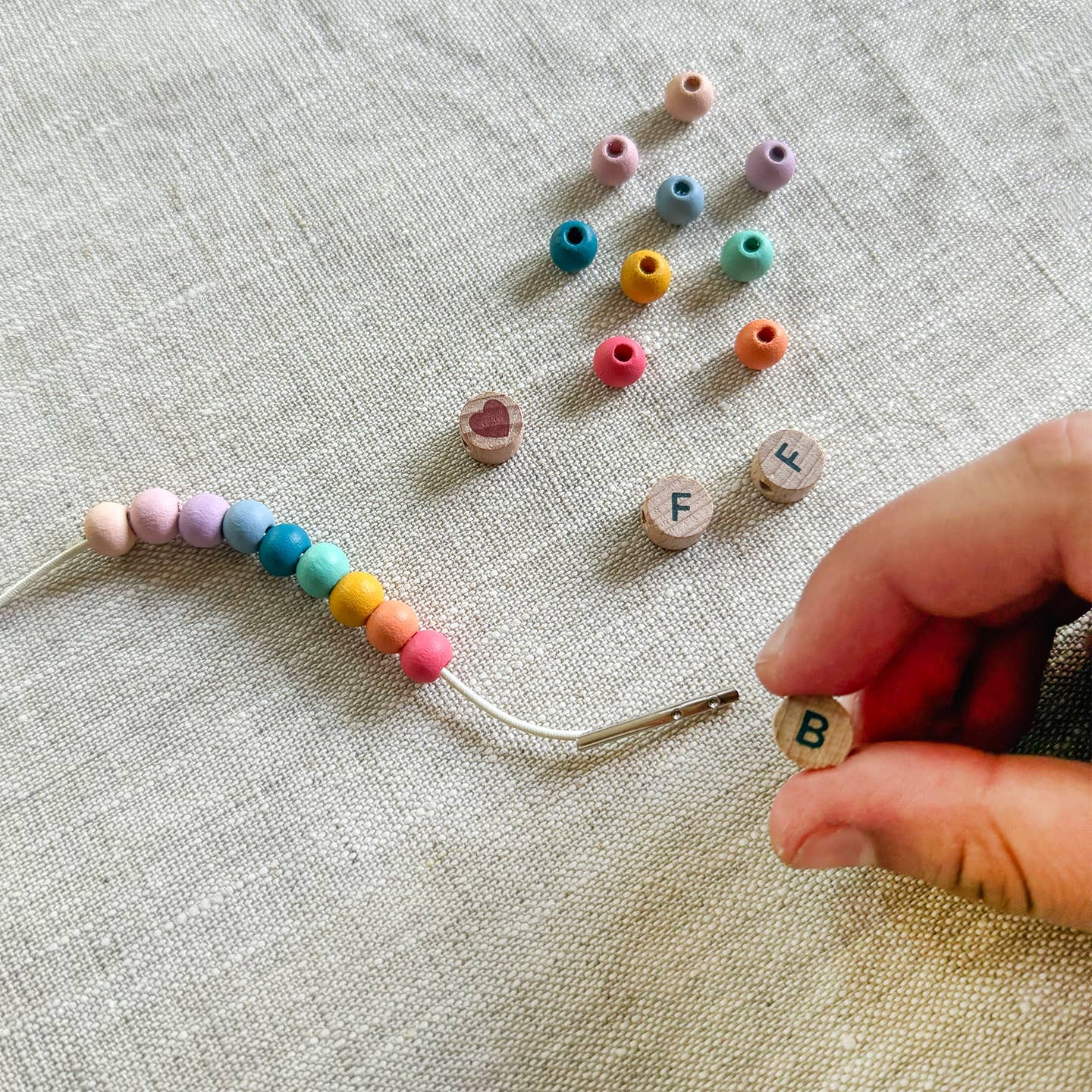 Colorful beads and letter beads on a textured fabric surface with a hand holding a needle.
