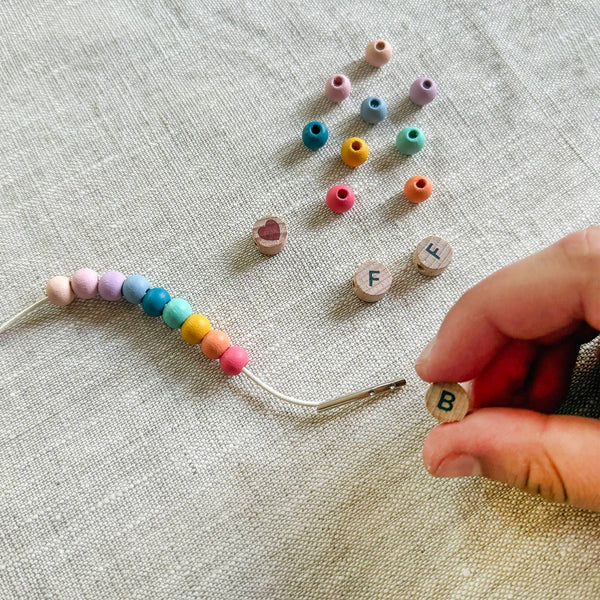 Colorful beads and letter beads on a textured fabric surface with a hand holding a needle.