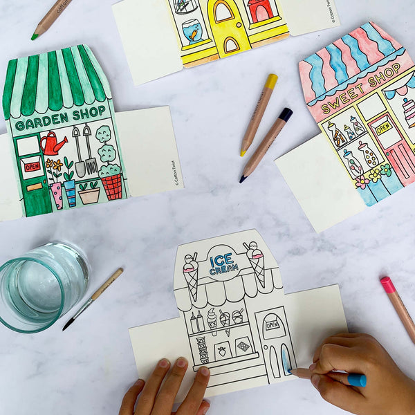 Coloring books with illustrations of shops on a table with coloring supplies.